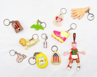 May include: A collection of vintage keychains with various designs, including a bird in a cage, a green pterodactyl, a baby, a miniature can, a running shoe, a hand, a bear with a heart, a worm, a cracked egg, and a patriotic design.