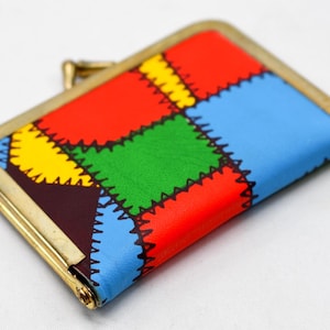 以下が含まれることがあります： 金色のフレームが付いた、カラフルな長方形の小銭入れ。赤、緑、黄、青のセクションで構成されたパッチワークデザインです。縫い目は黒い糸で縫われ、留め金は金色です。