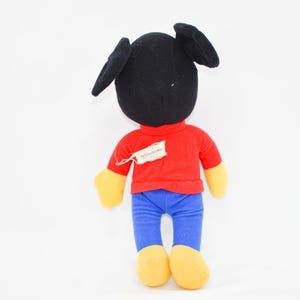 May include: A plush toy of a cartoon character, viewed from the back. The toy has a black head with large ears, a red shirt, blue pants, and yellow hands and feet. A tag is attached to the shirt.