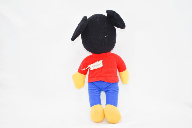 May include: A plush toy of a cartoon character, viewed from the back. The toy has a black head with large ears, a red shirt, blue pants, and yellow hands and feet. A tag is attached to the shirt.