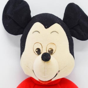 May include: A plush toy featuring the face of Mickey Mouse. The toy has large black ears, a cream-colored face, and a red shirt. The eyes are brown, and the nose is black. The toy is set against a white background.