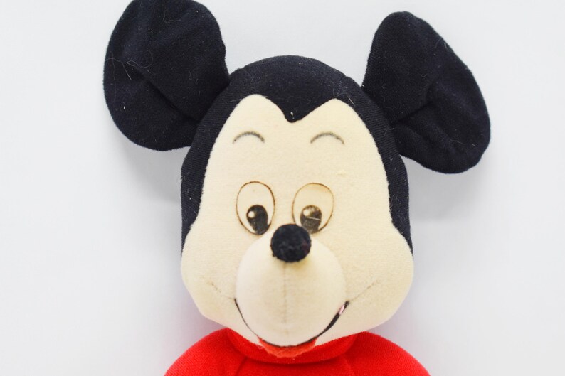 May include: A plush toy featuring the face of Mickey Mouse. The toy has large black ears, a cream-colored face, and a red shirt. The eyes are brown, and the nose is black. The toy is set against a white background.