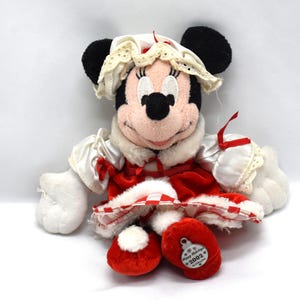 May include: A Minnie Mouse plush toy dressed in a red and white holiday outfit. The plush has black ears, a pink face, and is wearing a white bonnet with lace trim. The outfit includes a red velvet dress and matching red shoes. A tag on the shoe reads "Happy Holidays 2002."