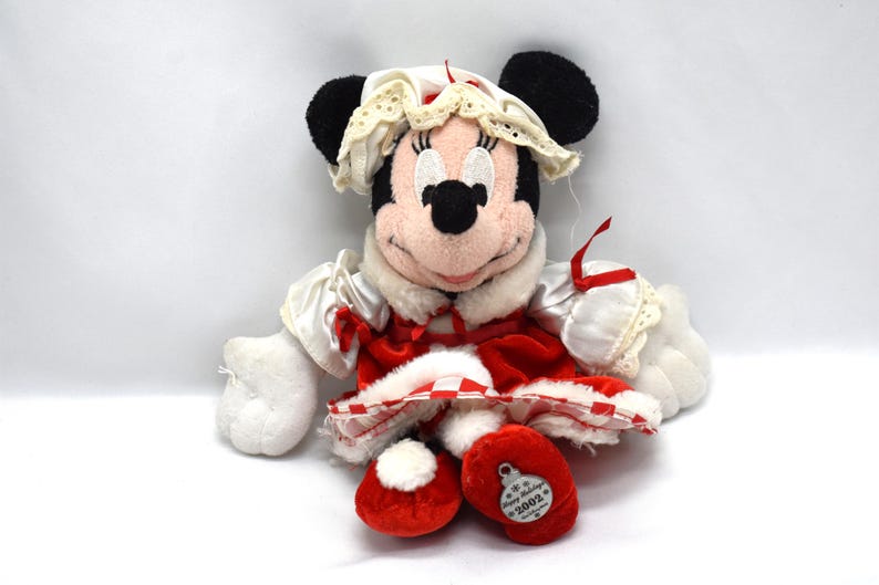 May include: A Minnie Mouse plush toy dressed in a red and white holiday outfit. The plush has black ears, a pink face, and is wearing a white bonnet with lace trim. The outfit includes a red velvet dress and matching red shoes. A tag on the shoe reads "Happy Holidays 2002."