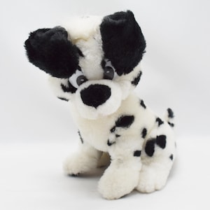 以下が含まれることがあります： 白地に黒い斑点のあるダルメシアン犬のぬいぐるみ。おもちゃは黒い耳、黒い鼻、大きくて丸い目をしています。ぬいぐるみは直立しています。
