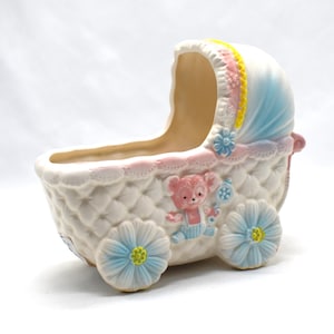 May include: A white ceramic baby carriage planter with a quilted pattern. It has a blue and pink canopy, floral details, and a pink teddy bear design. The wheels are light blue with yellow centres.