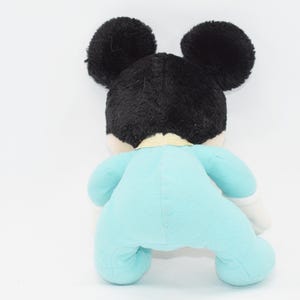 Op de afbeelding: Een pluche speelgoed van een bekende stripfiguur, van achteren gezien. Het speelgoed heeft zwarte oren en haar, een lichtblauwe onesie en witte handschoenen. Het speelgoed staat op een witte achtergrond.