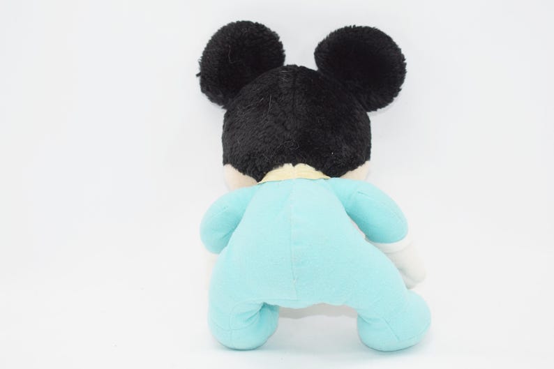 Op de afbeelding: Een pluche speelgoed van een bekende stripfiguur, van achteren gezien. Het speelgoed heeft zwarte oren en haar, een lichtblauwe onesie en witte handschoenen. Het speelgoed staat op een witte achtergrond.