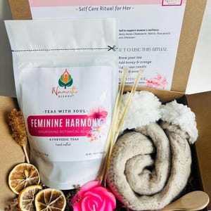 May include: A gift set in a cardboard box includes a bag of tea, a pink rose-shaped candle, a rolled beige scarf, a wooden spoon, and dried lemon slices. The bag of tea has the text "FEMININE HARMONY" and "TEAS WITH SOUL".