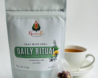 May include: A package of Daily Ritual Ayurvedic tea with a white teacup and saucer. The package has the text "Namaste Blends" and "Daily Ritual" with the words "Detox Cleanse Refresh". A small tea bag is in front of the package.