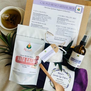 May include: A wellness gift box with a variety of self-care items. The box includes Vata Calm tea, a calming pillow mist, lavender eye pillows, and magnesium soak. A wooden spoon and a cup of tea are also visible.