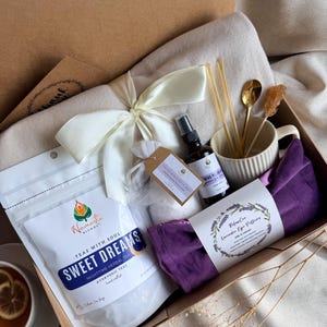 May include: A gift box containing a pouch of Namaste Blends Sweet Dreams herbal tea, a purple velvet eye pillow, a small spray bottle, a cream-colored ribbed mug with a gold spoon, a rock candy stirrer, and a white ribbon tied around a blanket.