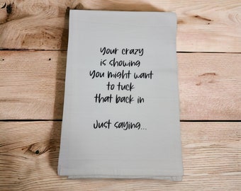 May include: A folded, light gray kitchen towel with the text "Your crazy is showing, You might want to tuck that back in, Just saying..." printed in black. The towel is on a wooden surface.