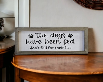 May include: A rustic wooden sign with a white background and black text that reads "the dogs have been fed don't fall for their lies". The sign has a distressed wood frame and paw prints on either side of the text.