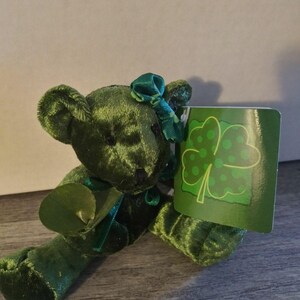 May include: A green plush teddy bear with a bow and a tag featuring a four-leaf clover design. The bear is made of a soft, velvety material and is holding a green tag with a shamrock illustration. The bear is a St. Patrick's Day themed item.