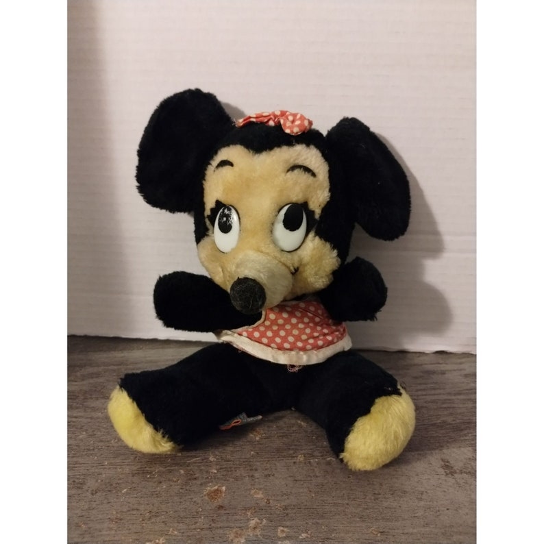 Puede incluir: Un juguete de peluche vintage de Minnie Mouse con pelaje negro, un vestido de lunares rojos y blancos y un lazo rosa. El juguete tiene ojos grandes y negros y una nariz negra.