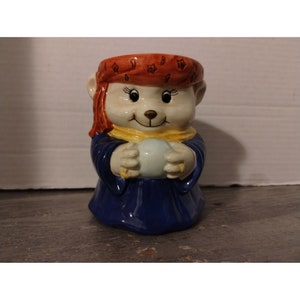 May include: A ceramic figurine of a bear-like character wearing a blue robe, yellow scarf, and a red head covering with star patterns. The character is holding a light blue ball. The figurine has a smiling face and is likely a decorative item.