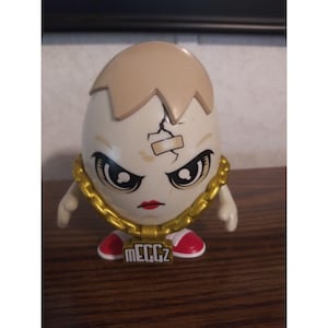 May include: A white egg-shaped figurine with a cracked head and a bandage. It has a stern face with large eyes, red lips, and a gold chain. The feet are red and white, and the text "MEGGZ" is on the chest.