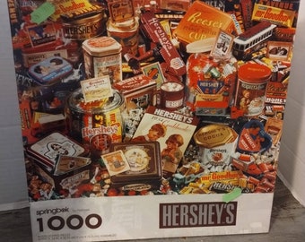 May include: A 1000-piece jigsaw puzzle featuring a colorful collage of Hershey's chocolate products, including candy bars, boxes, and tins. The puzzle box features the Hershey's logo and the text "Springbok by Hallmark".