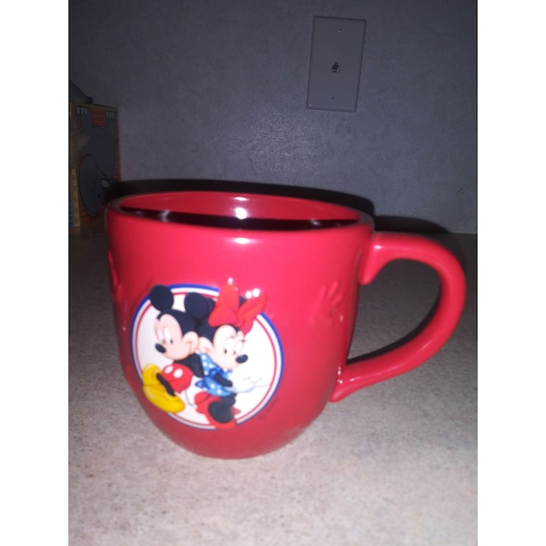 May include: Red ceramic mug with a round image of Mickey Mouse and Minnie Mouse in a circle. The image is on the front of the mug. The mug has a white handle.