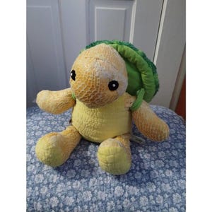 May include: A plush turtle toy with a yellow body and limbs, a green shell, and black eyes. The turtle is sitting on a blue floral patterned surface. The toy has a textured pattern on its head and limbs.