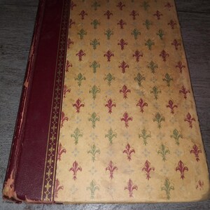 May include: A vintage book with a burgundy cover and a gold-colored spine. The cover features a repeating pattern of green and red fleur-de-lis on a cream background.