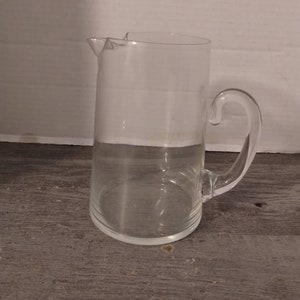 May include: A clear glass pitcher with a curved handle and a spout. The pitcher is cylindrical with a slightly wider base. The handle is attached to the side of the pitcher. The pitcher is sitting on a wooden surface.