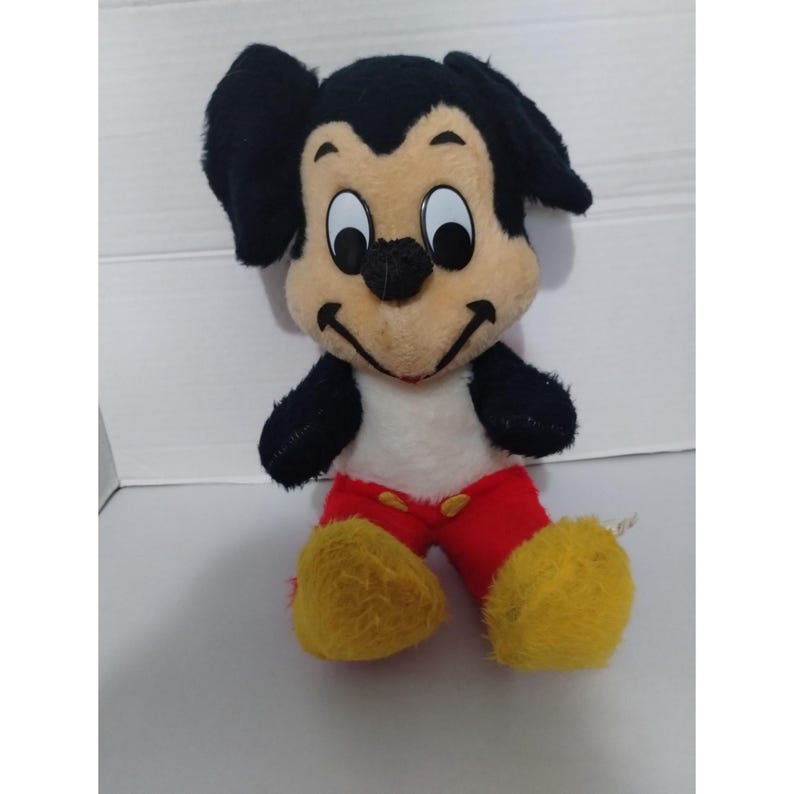 May include: A vintage plush Mickey Mouse toy with black ears, a black nose, and large eyes. The toy is wearing red pants with yellow shoes and a white shirt.