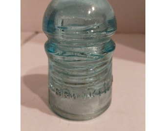 May include: A vintage, light blue glass insulator with a rounded top and a cylindrical base. The insulator has a series of horizontal ridges and the word "Brookfield" is embossed on the side. The glass has a slight aqua tint.
