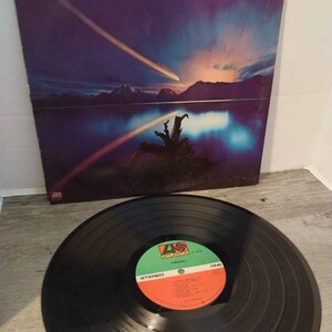 May include: A vinyl record album cover and record featuring the band Firefall. The cover art features a purple and blue sky with two shooting stars and a lake with a tree stump in the foreground. The record label is orange and green with the band name and album title.