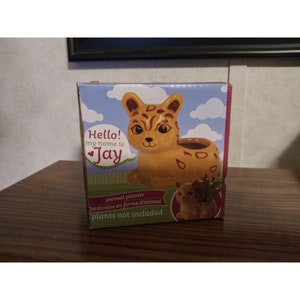 May include: A yellow and brown ceramic planter shaped like a cheetah with the words "Hello! My name is Jay" on the front. The planter is packaged in a box with a white background and blue clouds.