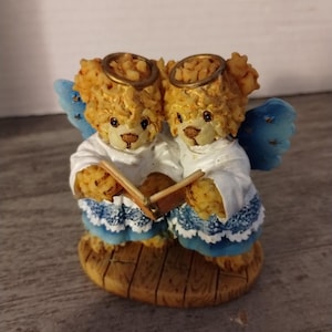 May include: Two teddy bears dressed as angels holding a book, with blue and white wings and gold halos. The bears are standing on a wooden base.