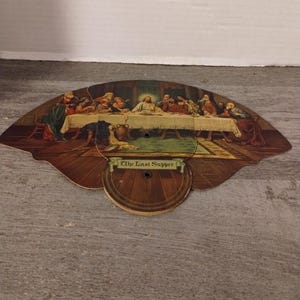 May include: A vintage hand fan depicting The Last Supper with a brown wooden frame and the words "The Last Supper" in a banner.