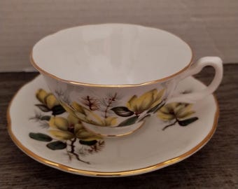 May include: A white teacup and saucer with a yellow floral pattern and gold trim. The cup and saucer are decorated with yellow flowers, green leaves, and brown branches.