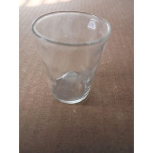 May include: A clear, small, empty glass with a slightly textured surface. The glass has a rounded bottom and a wide opening. The glass is designed to hold liquids and is suitable for various beverages.