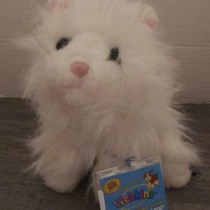 May include: A white plush toy cat with black eyes, a pink nose, and long whiskers. The toy has a tag that reads "Webkinz" and "Discover a virtual world!" The cat is sitting on a wooden surface.