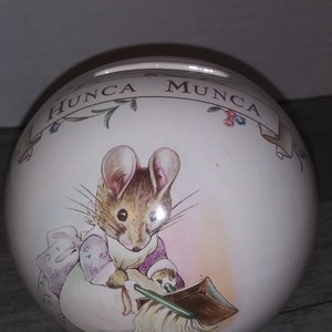 May include: A white ceramic piggy bank with a cartoon mouse sweeping. The bank has a floral design and the words "HUNCA MUNCA" written on it.