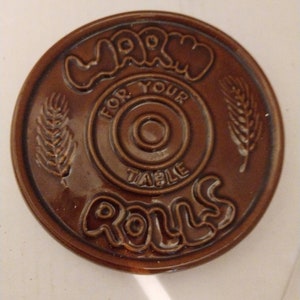 May include: Brown ceramic warm rolls holder with the text "Warm Rolls For Your Table" in a circle with wheat stalks on either side.