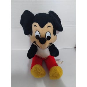 May include: A vintage plush Mickey Mouse toy with black ears, a black nose, and large white eyes. The toy is wearing red pants with yellow shoes and has a white belly.