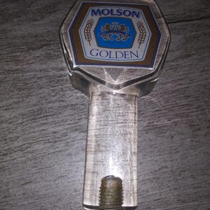 May include: A clear plastic beer tap handle with a gold and blue label that reads "Molson Golden".