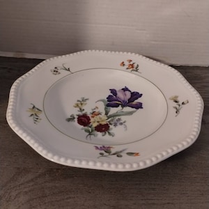 May include: White ceramic dinner plate with a floral design. The plate has a scalloped edge and a beaded rim. The floral design features a purple iris, red and yellow flowers, and green leaves.