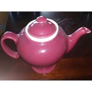 May include: A burgundy ceramic teapot with a white rim and lid. The teapot has a round body and a spout on the side.