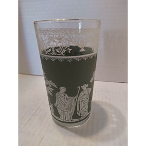 May include: A green glass tumbler with a white design of two figures in a garden setting. The glass has a gold rim and a white lace pattern around the top.