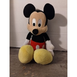 May include: A plush Mickey Mouse toy sitting with his legs crossed. He is wearing his signature red shorts with a white button and yellow shoes.