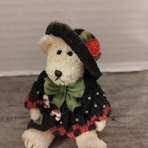 May include: A white plush teddy bear wearing a black and green sweater with a red and white candy cane design. The bear is wearing a black hat with a red flower and a green bow.