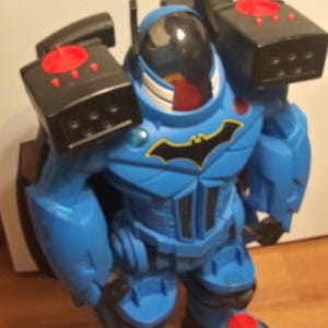 May include: Blue and black toy robot with a Batman logo on the chest. The robot has a clear dome head and red buttons on the arms.