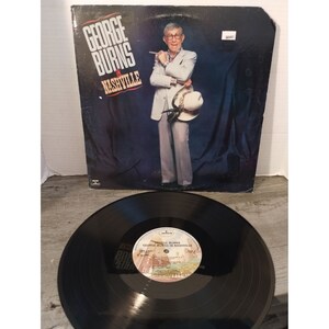 May include: A vinyl record album cover featuring comedian George Burns in a white suit and hat, with the title "George Burns in Nashville" in red and black lettering.