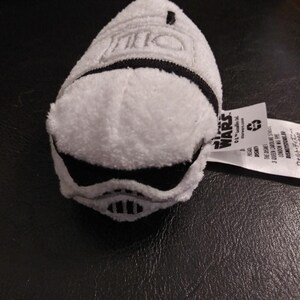 May include: A white plush toy shaped like a Stormtrooper helmet from Star Wars. The toy has black detailing for the visor and a black stripe around the base. The Star Wars logo is visible on a tag attached to the toy.