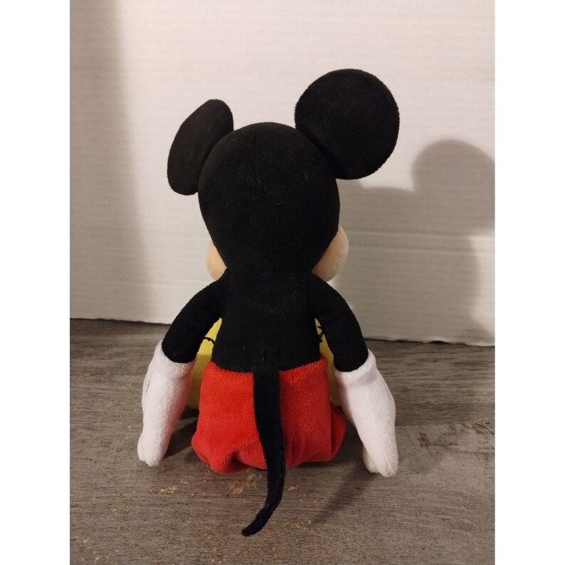 May include: A plush black Mickey Mouse toy with red shorts and white gloves. The toy is sitting on a yellow surface.