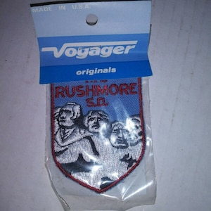 May include: A blue and red embroidered patch with the text "Mount Rushmore SD" and an image of the four presidents carved into the mountain.